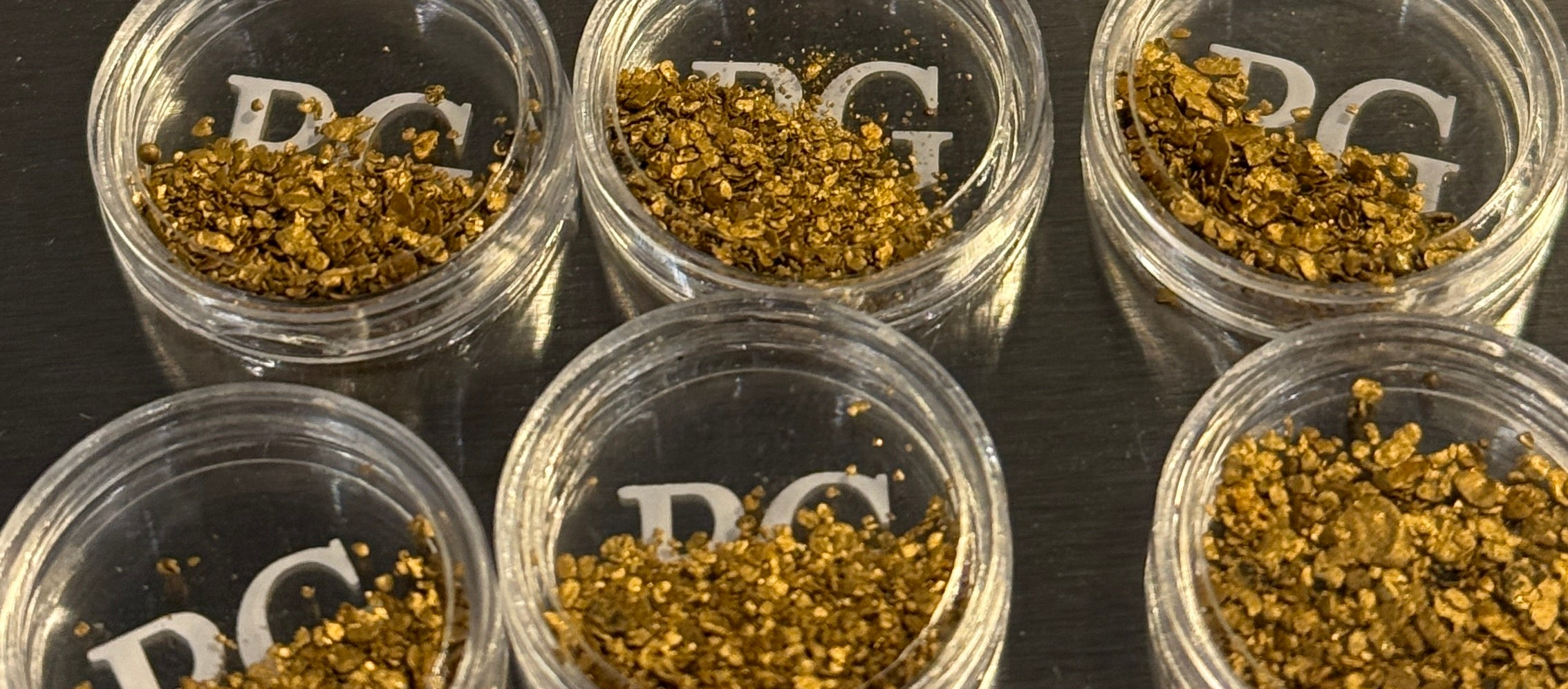 Small clear containers labeled 'PG' with raw gold on a dark surface.